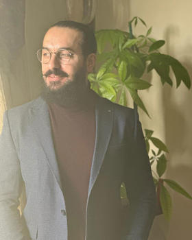 PhD student Nashaat Rasheed has a dark beard and wearing glasses, a dark suit, and turtleneck